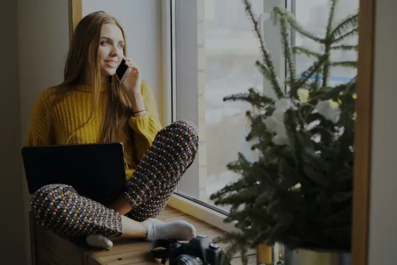 Une femme au téléphone près d’une fenêtre, ordinateur portable à côté d’elle.